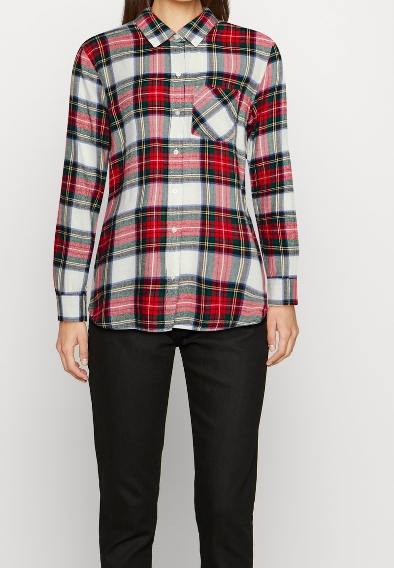 Femme portant une chemise à carreaux rouge, blanche et noire à boutons avec manches longues et un pantalon noir, debout devant un fond uni.