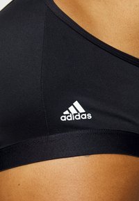 Crni atletski sportski grudnjak s glatkom teksturom, s bijelim Adidas logotipom na prednjoj strani, prikazujući detalje šivanja duž rubova.