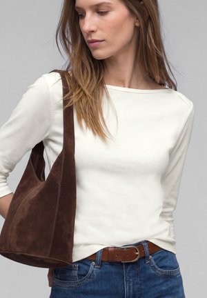 Frau trägt ein weißes langärmeliges Oberteil und blaue Jeans, führt eine große braune Wildleder-Schultertasche, schaut nach unten und zur Seite.