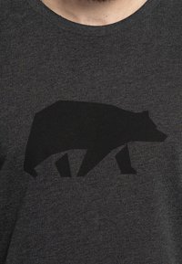 Camiseta gráfica negra con la silueta de un oso en negro sobre un tejido texturizado gris oscuro. Diseño de cuello redondo.