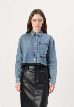 Denim shirt in light blue with a cropped design, button closure, and one chest pocket. Features a black lace trim and embroidered logo. Black leather skirt.