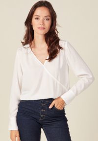Blusa blanca de manga larga con escote en forma de V, que presenta un diseño cruzado y una textura suave. Combinada con jeans azul oscuro con detalles de botones.