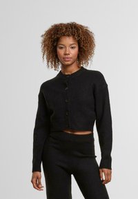 Fekete, rövidített kötött kardigán kerek nyakkivágással, gombos elülső résszel és hosszú ujjakkal. Puha textúra, amelyet egyszínű fekete kötött nadrággal párosítottak.