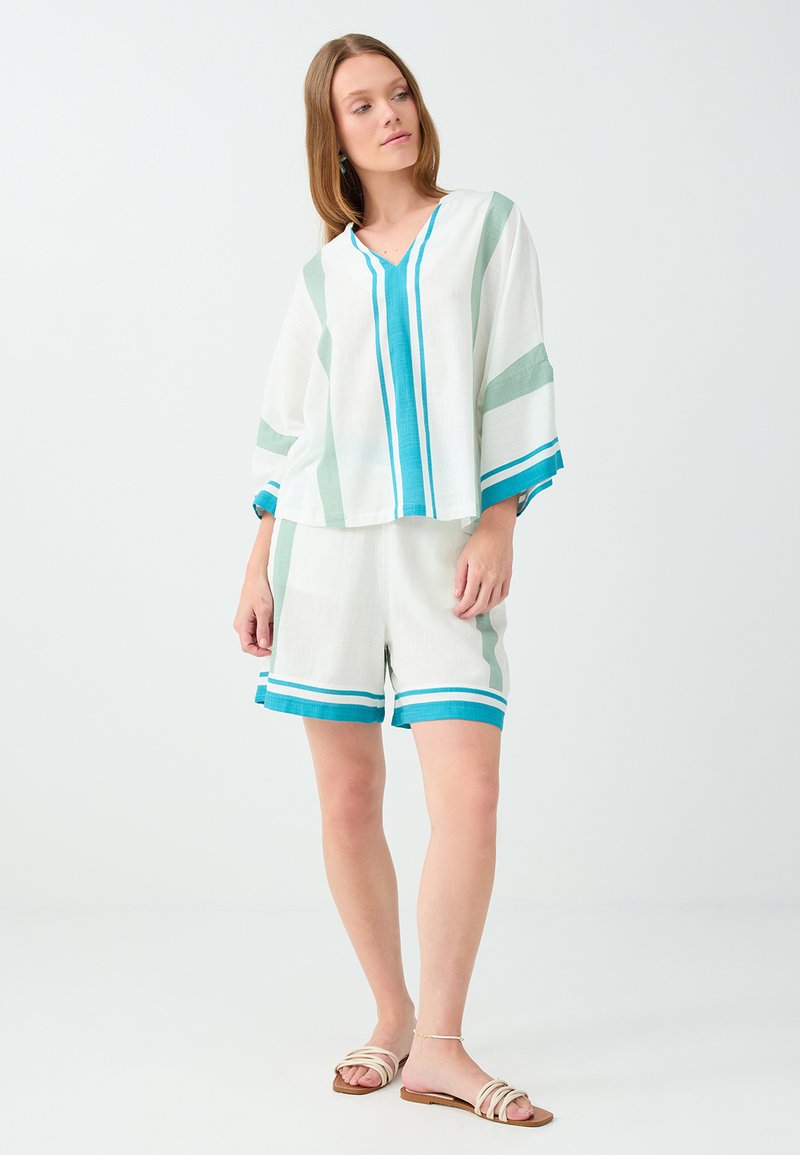 Femme portant un haut blanc à col en V et un short à rayures verticales bleues et vertes, debout, regardant sur le côté, portant des sandales beige.