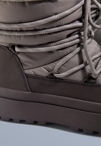 Botte d'hiver imperméable de couleur brun foncé avec une semelle en caoutchouc texturée, une tige grise matelassée et des lacets réfléchissants. Logo Primaloft visible.