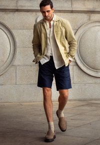 Homme marchant en plein air, portant une veste beige, une chemise blanche, un short bleu marine, des chaussettes beiges et des mocassins marron, devant un mur en pierre avec des détails circulaires.