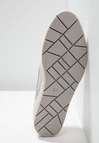 Semelle d’une chaussure gris clair avec un motif géométrique de la bande de roulement, indiquant la taille 37, positionnée verticalement contre un mur blanc.