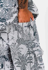 Mano con tatuaggio appoggiata nella tasca di pantaloni bianchi con dettagli di stampa botanica e animale nera, con il testo "Sixth June Paris".