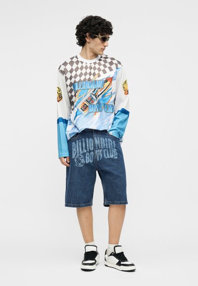 Άνδρας με γυαλιά ηλίου, πουκάμισο γραφικού τύπου με μακριά μανίκια που γράφει "Billionaire Boys Club", denim σορτς με το ίδιο κείμενο και ασπρόμαυρα αθλητικά παπούτσια.