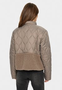 Quiltad beige jacka med en texturerad brun fleecepanel på nedre delen, med krage och lång ärm.
