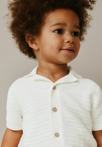 Vit texturerad kortärmad skjorta med krage och fyra ljusbruna knappar. Tyget har en ribbad design.