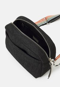 Sac bandoulière en tissu noir avec fermeture éclair ouverte montrant une poche intérieure, avec une sangle multicolore réglable et une boucle en métal.
