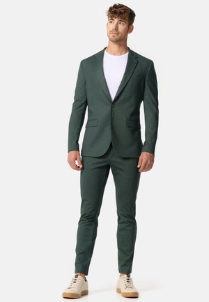 Green textured suit with a notched lapel, fitted design, paired with a white t-shirt and light beige sneakers. Features two pockets on the blazer.