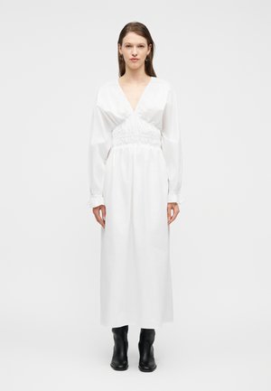 Donna con un lungo abito bianco con scollo a V profondo e maniche lunghe, abbinato a stivali neri fino a metà polpaccio, in piedi davanti a uno sfondo semplice.