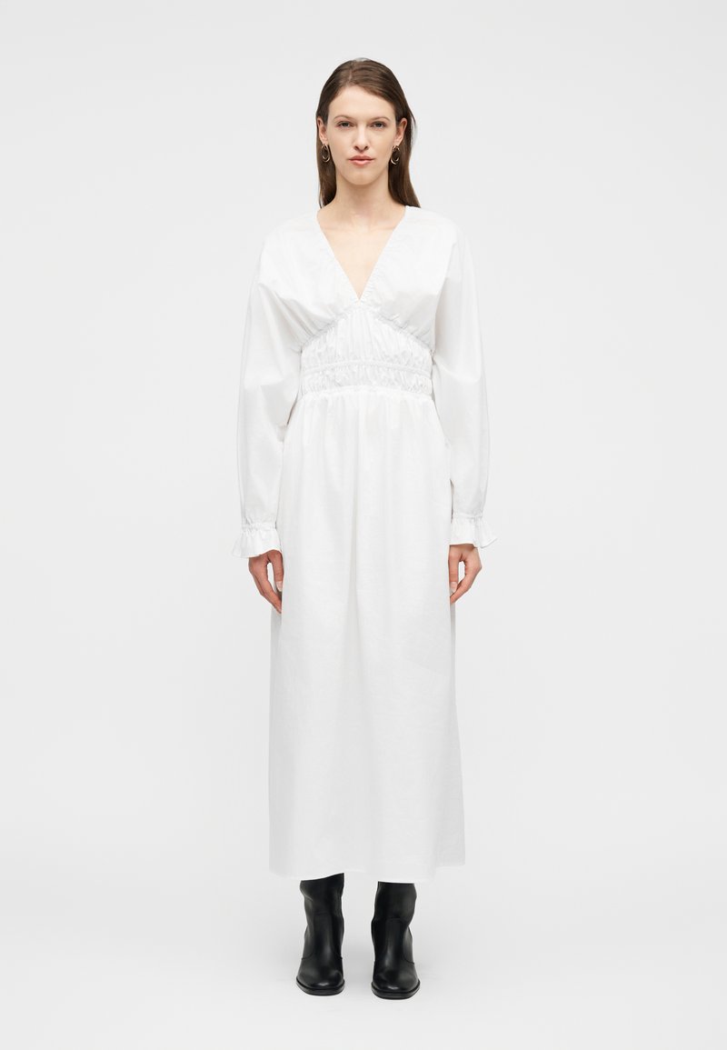 Γυναίκα με μακρύ λευκό φόρεμα με βαθύ V-λαιμόκοψη και μακριά μανίκια, συνδυασμένο με μαύρα μπότες μέχρι τη μέση της γάμπας, που στέκεται μπροστά σε απλό φόντο.