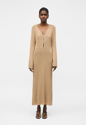 Frau trägt ein langes beiges geripptes Strickkleid mit langen Ärmeln, einem V-Ausschnitt mit zwei Schlüssellochausschnitten und schwarzen offenen Sandaletten mit Absatz.