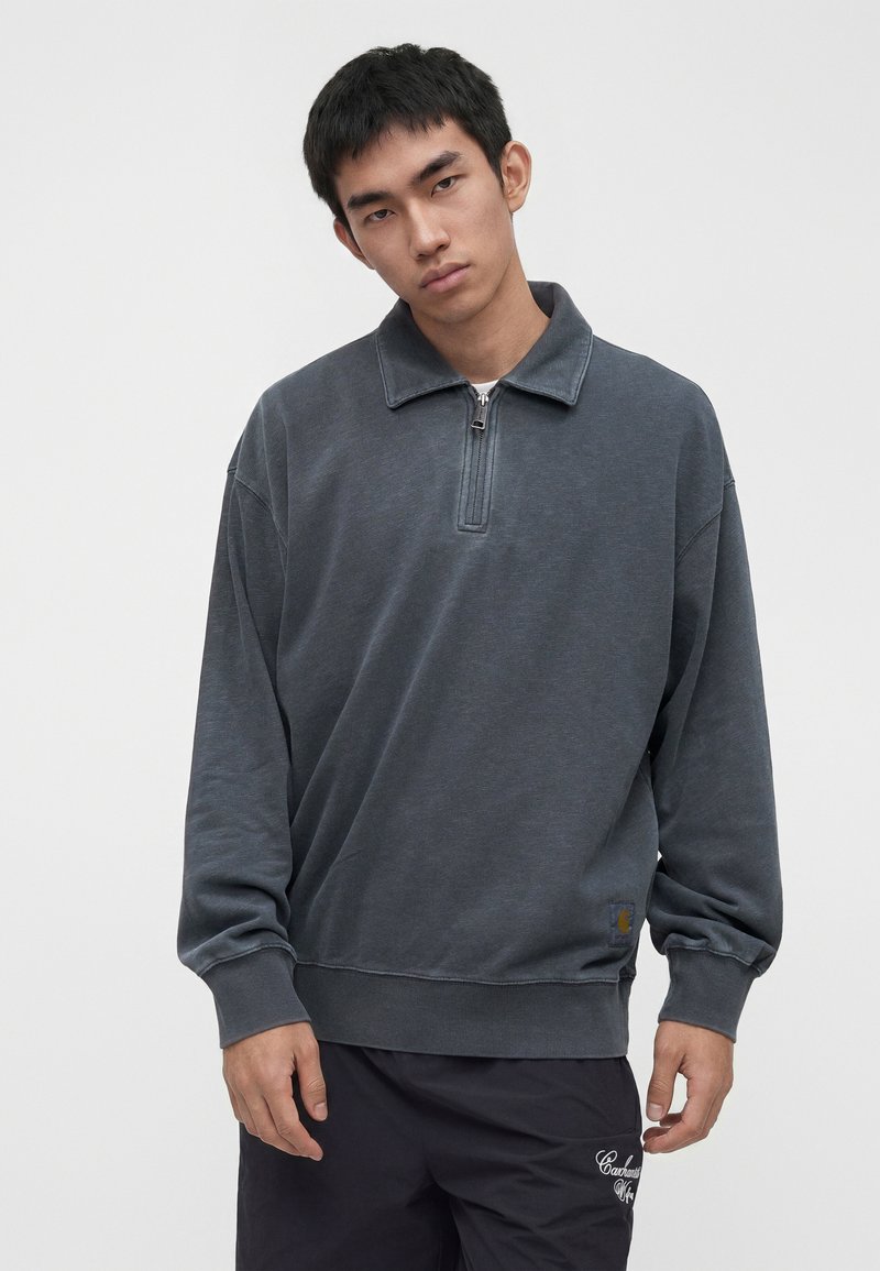 Young man wearing a dark grey zip-up collared sweatshirt and black pants with white embroidered text on the upper thigh.