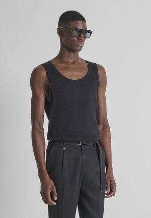 Man draagt een zwart mouwloos shirt, zwarte broek met riem en donkere zonnebril, staand tegen een effen grijze achtergrond.