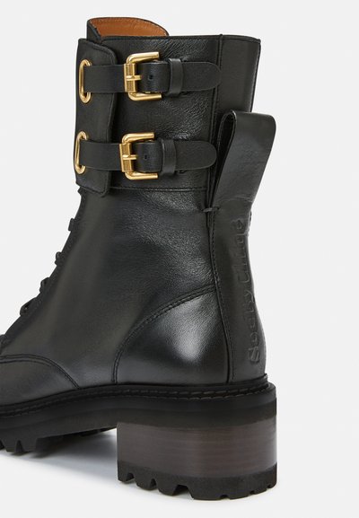 Black leather ankle boot with gold buckles, lace-up front, textured surface, and chunky wooden heel. Side features embossed branding detail.
