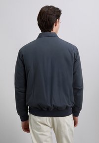 Man wearing a dark blue bomber jacket and light beige pants, standing with his back to the camera against a plain light gray background.