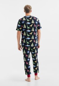 Jonge man in zwarte pyjama's met kleurrijke dinosaurussen en versierde kerstboom patronen, staande op blote voeten, achterkant zichtbaar.