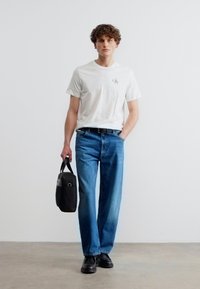 Άνδρας με λευκό T-shirt Calvin Klein, μπλε τζιν, μαύρα παπούτσια, κρατώντας μια μαύρη δερμάτινη τσάντα με το ένα χέρι, στέκεται μπροστά σε μια λευκή απλή τοίχο.