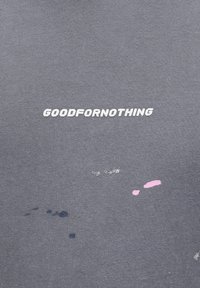 T-shirt in tessuto grigio con la scritta "GOODFORNOTHING" in bianco. Include macchie di vernice blu e rosa. Texture morbida.