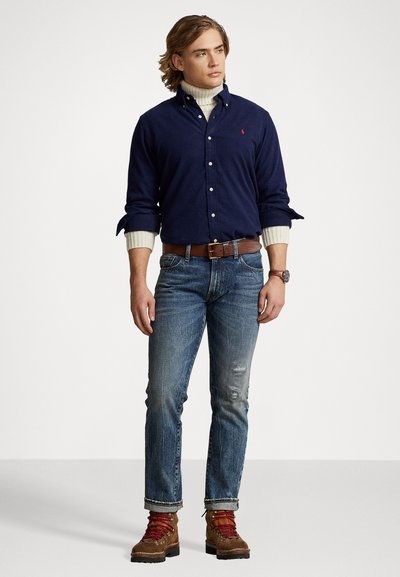 Camisa azul-marinho de botão, gola alta creme, jeans azuis desfiados, cinto castanho e botas castanhas com cordões vermelhos; modelo encosta-se a um fundo neutro.