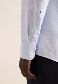 Camicia a maniche lunghe azzurro chiaro con motivo a quadretti, polsini con bottoni e un piccolo ricamo a forma di rosa vicino al polsino.