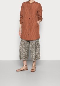 Brun, quiltet lang jakke med rund krave, sorte knapper og sidelommer, parret med blomstrede vide bukser og metalliske sandaler.