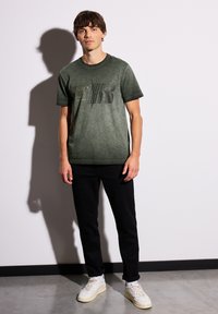 Zelené melírované tričko s černým textem a grafickým designem, v kombinaci s černými kalhotami a bílými teniskami na jednobarevném pozadí.