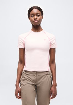 Γυναίκα με σκουρόχρωμο δέρμα και μαλλιά φοράει ανοιχτό ροζ κοντομάνικο πουκάμισο και μπεζ παντελόνι, στέκεται μπροστά σε απλό λευκό φόντο.