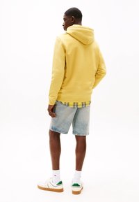 Felpa gialla con una trama morbida, abbinata a una camicia a quadri gialla e nera, pantaloni shorts in denim azzurro chiaro e sneakers bianche con accenti verdi.
