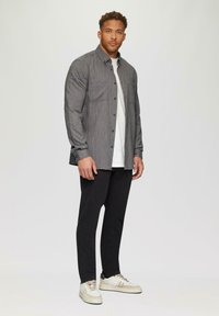 Grauhemd mit langen Ärmeln, zwei Brusttaschen, über einem weißen T-Shirt getragen; schwarze Hosen und cremefarbene Sneakers vervollständigen das Outfit.