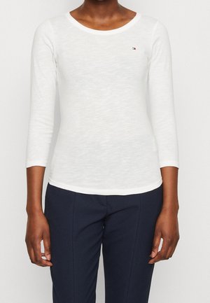 Haut blanc slim fit à manches longues avec encolure ronde, porté avec un pantalon foncé texturé, montrant le haut du corps et les bras.