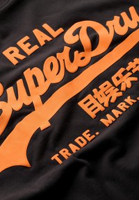 Sudadera negra con texto en relieve en color naranja que dice "REAL SuperDry", con acentos adicionales en naranja y una textura suave.