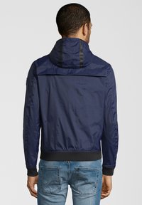Uomo che indossa una giacca a cappuccio blu navy e jeans blu, visto da dietro contro uno sfondo grigio chiaro uniforme.
