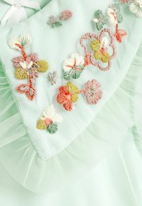 Tissu délicat vert pastel avec des motifs floraux brodés en blanc, rose, orange et vert, rehaussé par de douces bordures en tulle froncé.