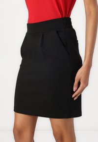 Black pencil skirt with pleats, elastic waistband, and two side pockets. Made of smooth fabric, offering a fitted silhouette.