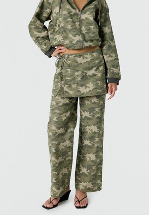 Nő zöld terepmintás cipzáras kapucnis pulóverben, hozzá illő, nadrág felett viselt átlapolt szoknyában és fekete ék-szandálban, keze a csípőjén áll.