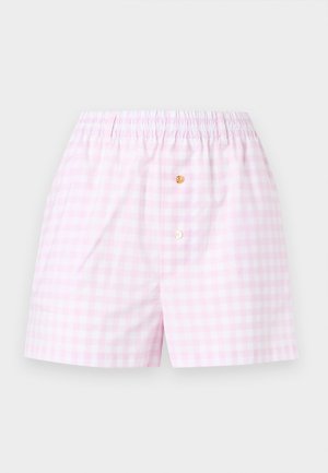 Γυναίκα στέκεται φορώντας λευκό μπλουζάκι Moschino, ανοιχτό ροζ σορτς και μαύρα σανδάλια με ψηλό τακούνι σε λευκό φόντο.