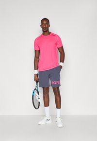 Αθλητική ενδυμασία που περιλαμβάνει ένα ροζ μπλούζα που απορροφά την υγρασία και γκρι σορτς με το λογότυπο "BORG". Περιλαμβάνει λευκές κάλτσες και αθλητικά παπούτσια, κρατώντας ένα ρακέτα.