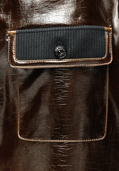 Gros plan sur la poche d'une veste en cuir marron brillant avec un rabat en tissu noir maintenu par un bouton-pression noir.