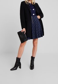 Zwarte jas over een marineblauwe top met stippen, gecombineerd met een geruite rok. Een zwart gestructureerde clutch en enkellaarsjes met hak maken de look compleet.