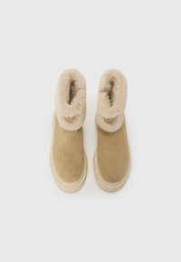 Bottes chelsea beiges avec un col rabattable doux et pelucheux, en matériau suédé et une semelle texturée. Accentué par un emblème décoratif sur le côté.