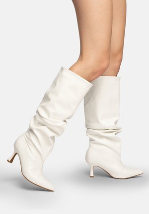 Nogi w białych luźnych, sięgających kolan skórzanych kozakach z ostrym noskiem i niskim obcasem na jasnym tle.