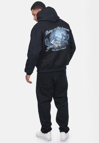 Zwarte oversized hoodie met een grafisch ontwerp van vogels en bergen op de achterkant, met lichtblauwe letters. Gecombineerd met donkere broeken en witte sneakers.