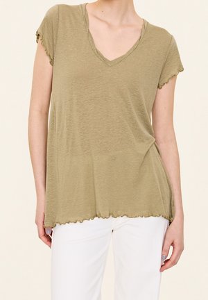 Kobieta ubrana w jasnooliwkowy top z krótkim rękawem i dekoltem w serek, z falbankowym brzegiem i mankietami na rękawach, w połączeniu z białymi spodniami.