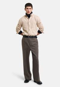 Camisa beige de botones sobre un cuello de tortuga azul marino, combinada con pantalones amplios de tweed marrón y un cinturón negro. Unos zapatos negros completan el conjunto.