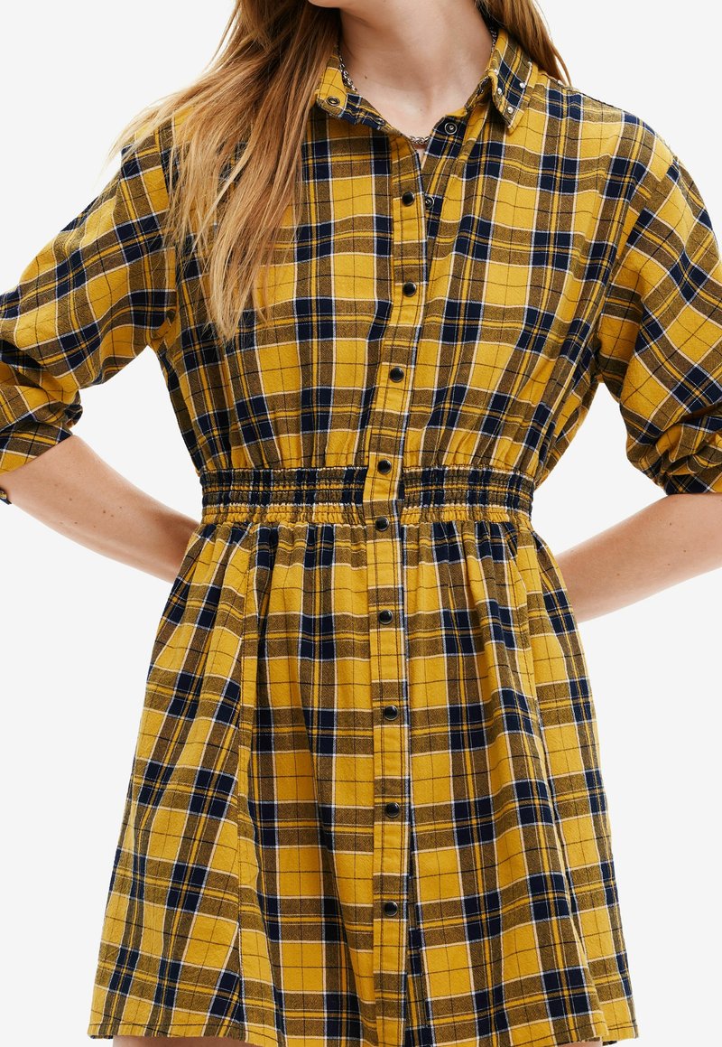 Donna che indossa un abito a quadri giallo e blu navy con vita elasticizzata e maniche arrotolate.